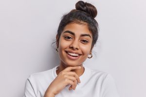 Portrait of tender charming woman touches chin gently smiles broadly has healthy skin being in good mood glad to pose for making photo isolated over white background. Happy human emotions concept
